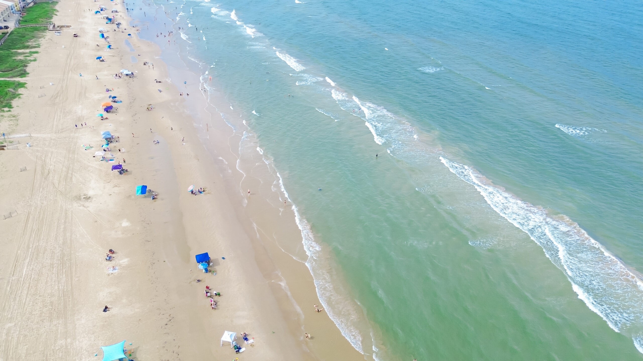Sandy shoreline, calm waves emerald water along row of beach condos, hotels, resort in South Beach, South Padre Island, exquisite barrier Gulf Coast of Texas, people enjoy beach activities aerial. USA